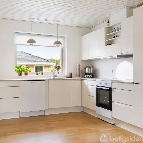 Et lyst, moderne køkken med hvide skabe og en ovn. To lamper hænger over bordpladen. Vindue med udsigt til et hus. Plante og køkkenmaskine ved vinduet.