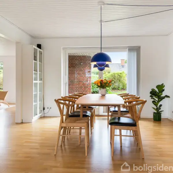 Spisebord med otte stole og en blå loftslampe, blomsterdekoration i midten. Rummelig, lys stue med trægulv, stor kunstplakat på væggen, åbning til stue med siddepladser.