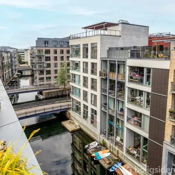 Lejlighedskompleks med store vinduer og altaner ligger ved en kanal. Broer forbinder bygningerne, hvor både ligger fortøjet. Tagterrasse til venstre med planter. Urban miljø.