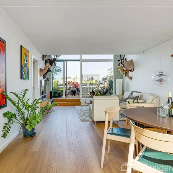 Et moderne stueområde med et rundt spisebord, stole og planter. Kunstværker og dyrehoveder dekorerer væggene. I baggrunden ses en sofa ved et stort vinduesparti med udsigt til en altan.