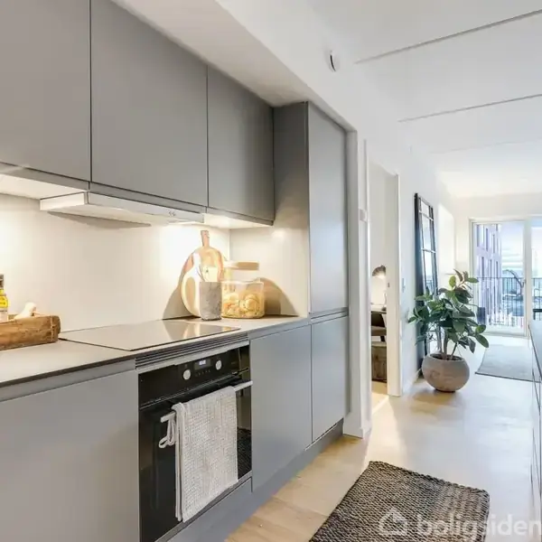 Et moderne køkken i neutrale farver har en kogeplade og ovn indbygget i et skab. En plante og flasker står på bordet. Spiseområdet ses i baggrunden.