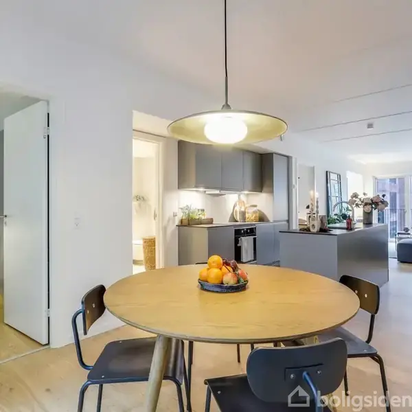 Et rundt spisebord med frugtskål står i en moderne stue-køkken kombination. Lyse trægulve strækker sig gennem rummet, der er indrettet med grå sofaer og store vinduer.