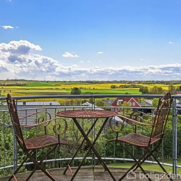 Et rundt cafébord med to stole står på en balkon. De vender ud mod en udsigt af marker og huse under en blå himmel med spredte skyer.