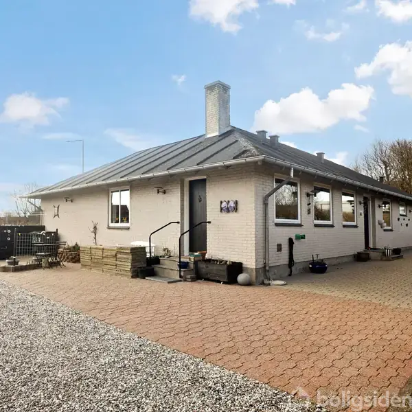 Huset er et hvidt murstenshus med et gråt sadeltag. Foran huset ligger en flisebelagt indkørsel og grusbelagt område, omgivet af hække og buske under en blå himmel med skyer.