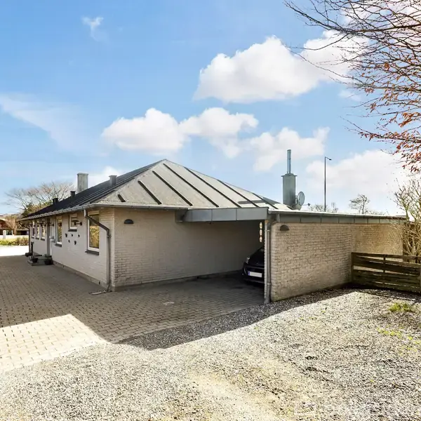 Hus med fladt tag står stille i en solrig indkørsel med stenfliser, omgivet af hække og træer. En bil er delvist parkeret under en carport.