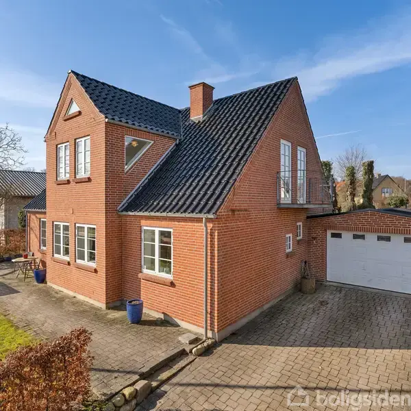 Rødt murstenshus med sort tegltag står stille ved solbeskinnet gårdsplads. Garage er til højre. Omkranset af hæk og græsplæne i et roligt boligkvarter.