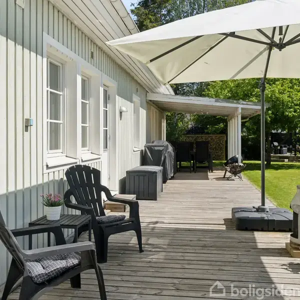Træterrasse med sorte stole og et bord. En stor parasol giver skygge, og en dekorativ mælkekande står ved siden af blomster. Græsplæne omgiver området. Ingen tekst til stede.