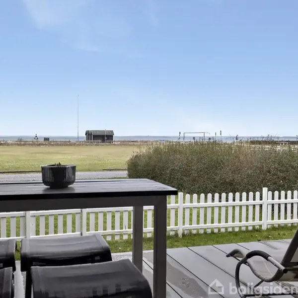 Et træbord med stol på terrasse, udsigt over græsplæne mod havet. En hvid stakithegner terrasseområdet, solen skinner klart.