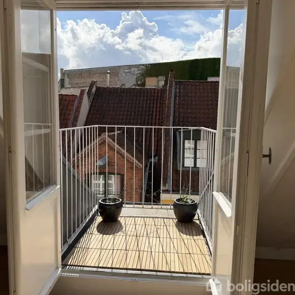En lille balkon med to potteplanter står åben gennem franske døre. I baggrunden ses hustage og en blå skyet himmel.