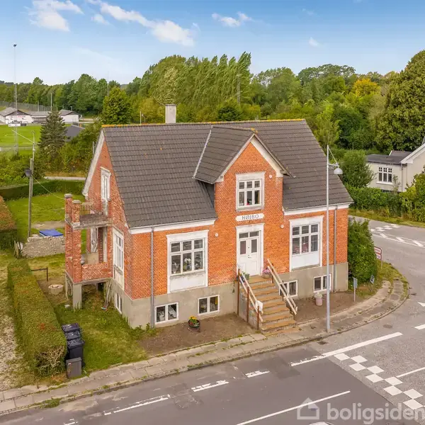 Et rødt murstenshus med hvide detaljer står stille ved en vej i en grøn, landlig omgivelser. Huset har en trappe op til hoveddøren og parkering ved siden af.