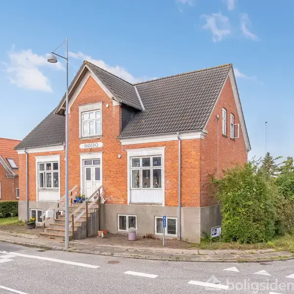 Rødt murstenshus med hvide vinduer og trappe på forsiden står ved vejen under en blå himmel. Skilte læses "MIDDELVEJ". Træer og andre bygninger i baggrunden.