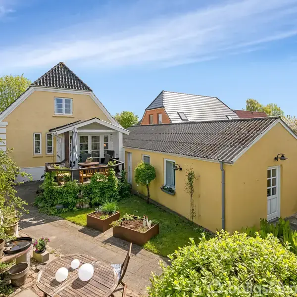 Gulmurstenshus med veranda og to stuer forbundet af en terrasse; omgivet af en frodig have med blomsterkrukker og havemøbler under en blå himmel.
