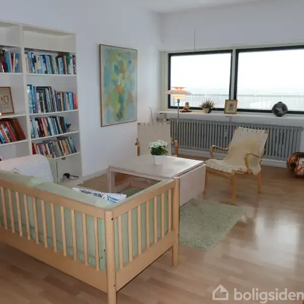 Sofa og lænestol står i en stue med store vinduer. Der er en fyldt bogreol til venstre, et sofabord i midten og diverse dekorationer, herunder kunstværk og planter.