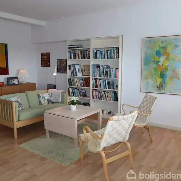En hyggelig dagligstue med en grøn sofa og to lænestole omkring et sofabord, omgivet af en bogreol fyldt med bøger og farverig kunst på væggene.