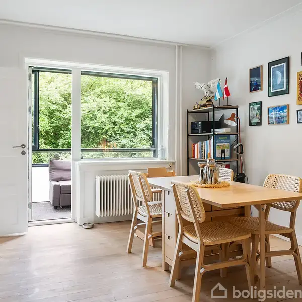Spisebord med fire stole står i et lyst rum. Bordet er dækket med glasflasker, og en hylde med bøger og dekorative genstande er langs væggen. En åben dør leder til terrasse med udsigt til grønt areal.