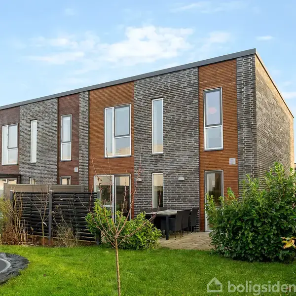 Rækkehus står stille i en haveomgivelse; huset har en moderne facade af mursten og træpaneler med store vinduer, mens en græsplæne med buske omgiver det.