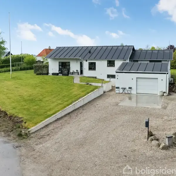Hvidt hus med sort tag står på en stor grusindkørsel, omgivet af en velplejet grøn græsplæne; træer og buske ses i baggrunden.