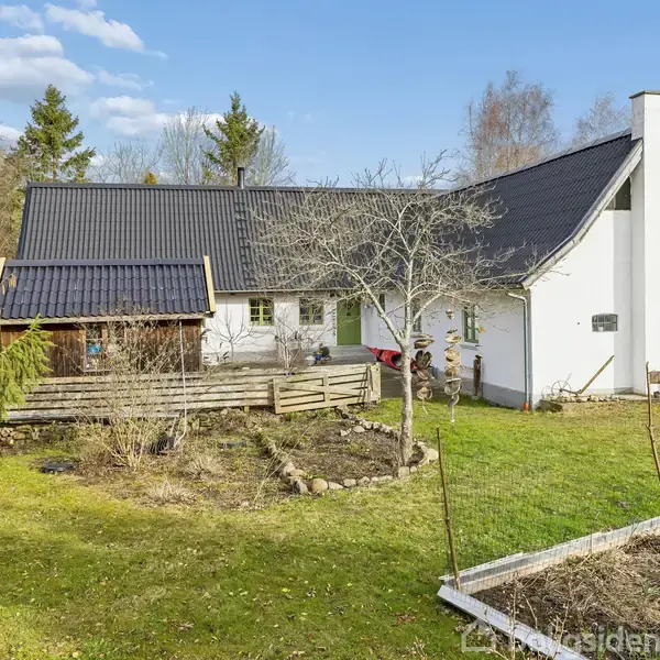 Hvidt hus med sort tag står i en have med græsplæne og blomsterbede. Der er træer i baggrunden, og det er solrigt med blå himmel og skyer.