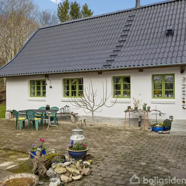 Hvidt hus med grå tag og grønne vinduer står stille. Der er havemøbler og planter på en flisebelagt gårdsplads omgivet af grønne træer. Grøn dør til højre.