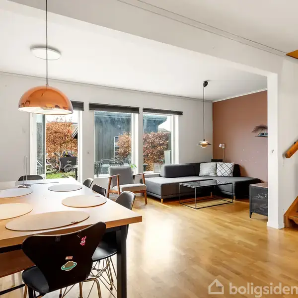 Spisebord med dækkeservietter står i en stue med lysindfald fra store vinduer. Sofa og trapper tilfører komfort og forbindelse til det øvrige hus i moderne omgivelser.