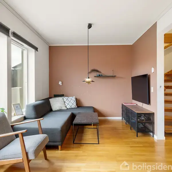 En stue med en grå hjørnesofa står op ad en brun væg. Et lille bord og en stol i træ ved siden af. En trappe fører opad til højre.