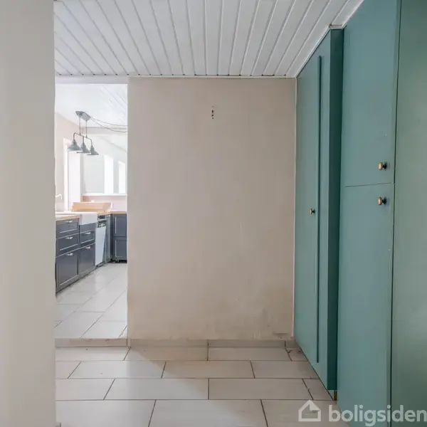 Gang med beige fliser og hvidt loft. Grønne skabe til højre, åbner mod køkken med mørkegrå skabe og hængelamper i baggrunden.