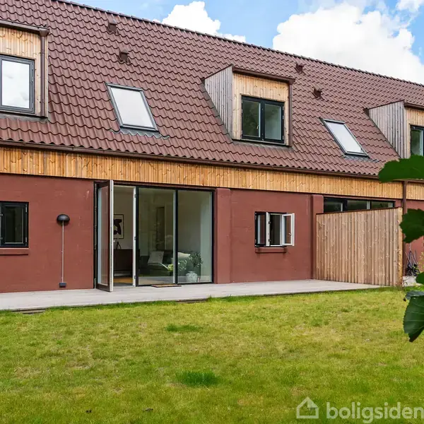 Hus med rødt tag og flere kvistvinduer. Store glasskydedøre åbner sig mod haven. Græsplænen foran huset er velholdt.