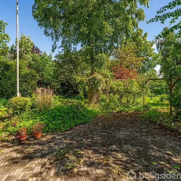 En brolagt terrasse med to tomme plantekrukker står ved siden af et grønt haveområde. Træer og buske omgiver pladsen, mens en træpergola er synlig til højre.