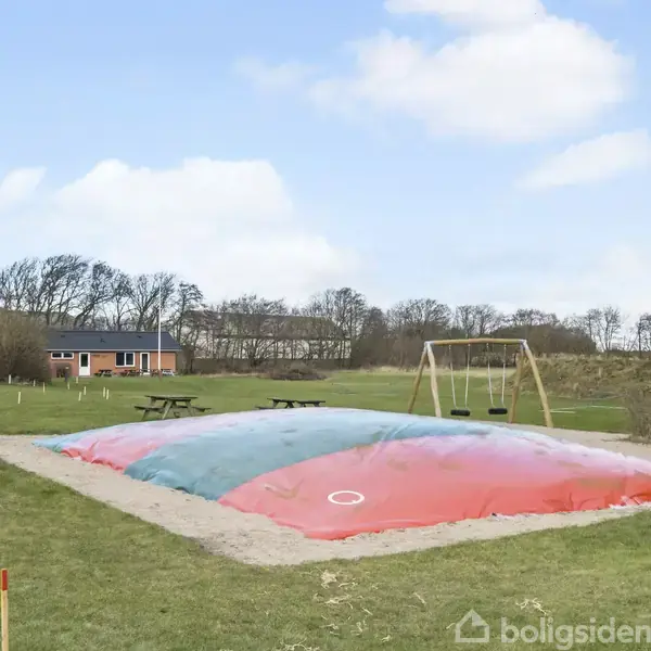 Hoppepude er oppustet i en legeplads omgivet af græsplæne og træer; der er gynger ved siden af, og et picnicbord står i nærheden.