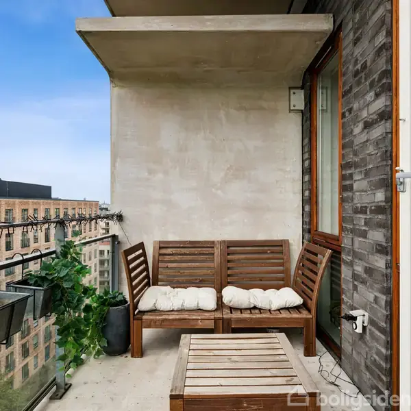 Balkonen har træmøbler med puder, placeret ved en murstensvæg. Den moderne byudsigt inkluderer høje bygninger. Grønne planter er arrangeret langs altanens glasrækværk.