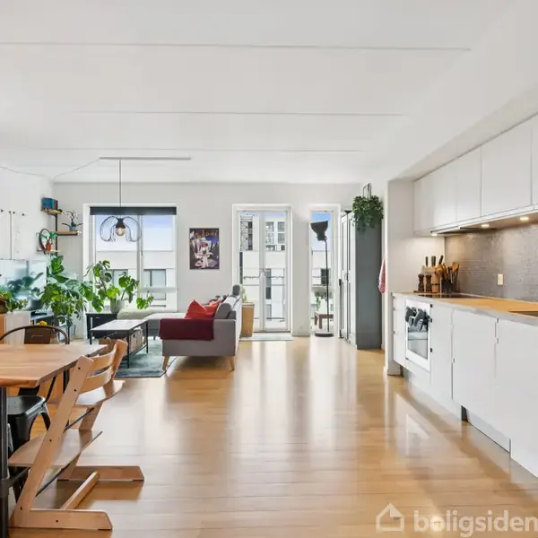 Lyst køkken-alrum med hvide skabe og træbord i forgrunden; sofaområde med rødt tæppe og grønne planter i baggrunden. Store vinduer tillader naturligt lysindfald.