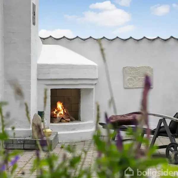 Udendørs pejs brænder lystigt ved siden af en sort liggestol i en gårdhave omgivet af hvide vægge og blomster, under en blå himmel med spredte skyer.