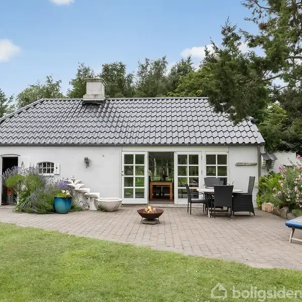 Hvidt hus med gråt tag står stille i en frodig have. På terrassen er der havemøbler og blomsterkrukker. Græsplæne og træer omslutter området, hvilket skaber en hyggelig atmosfære.