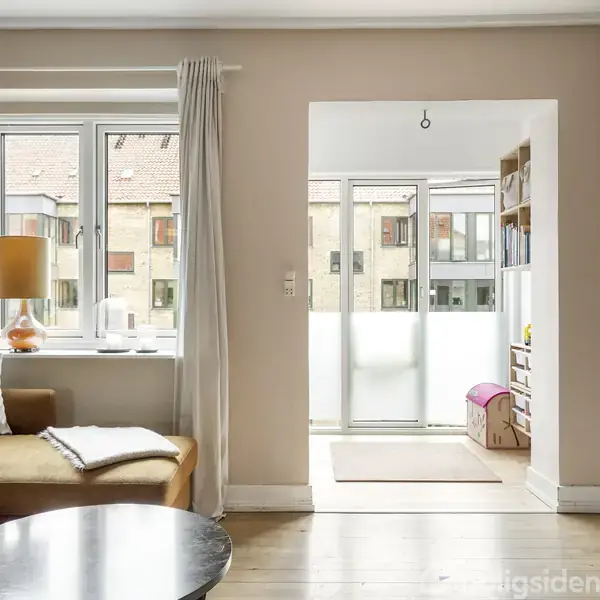 Stue med beige sofa og hvidt tæppe ved et rundt sofabord; vindue med gardiner og lampe på højre side. Åben væg fører til lysere rum med glasdør og hylder.