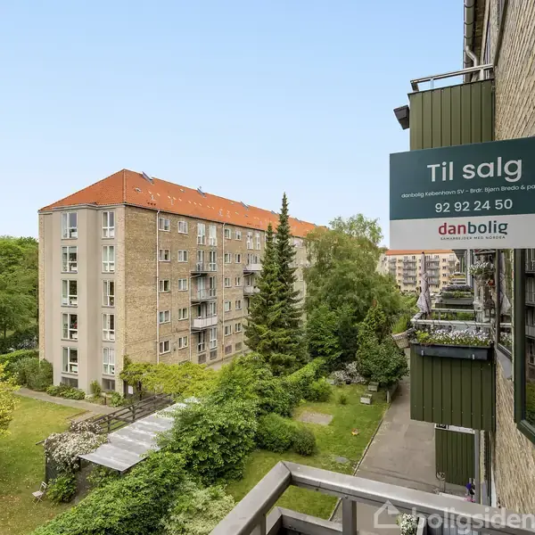 En lejlighedsbygning har et "Til salg" skilt hængende på facaden, med tekst: "Til salg, danbolig, 92 92 24 50". Udsigt fra en balkon viser grønne områder og nabobygninger.