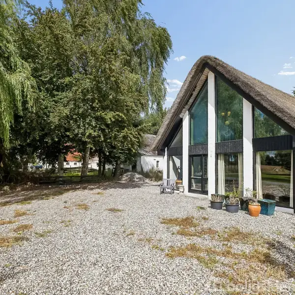 Et hus med stråtag og store glasvinduer står i en gårdsplads dækket af grus. Flere blomsterpotter står på terrassen. Omkring huset ses træer og en blå himmel.