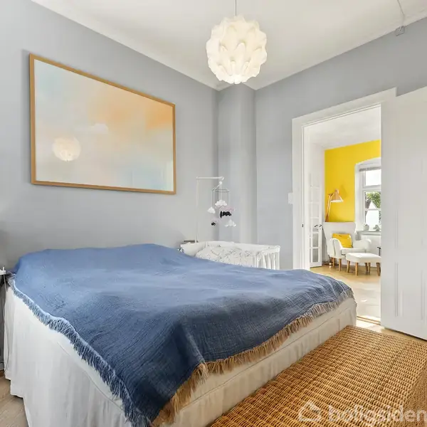 Soveværelse med en seng dækket af et blåt sengetæppe, omgivet af lyseblå vægge. En åben dør viser en gul væg i det tilstødende rum. En moderne lampe hænger fra loftet.