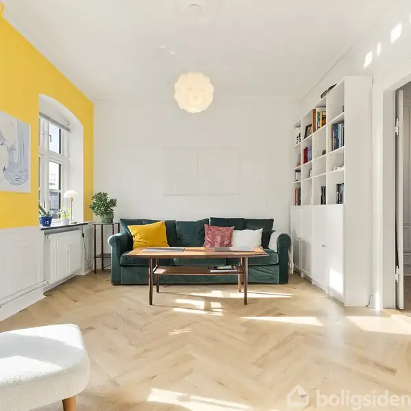 En stue med en grøn sofa og farverige puder, placeret foran en hvid reol. Rummet har gule vægge, lyst trægulv, og store vinduer. En lampe hænger centralt fra loftet.