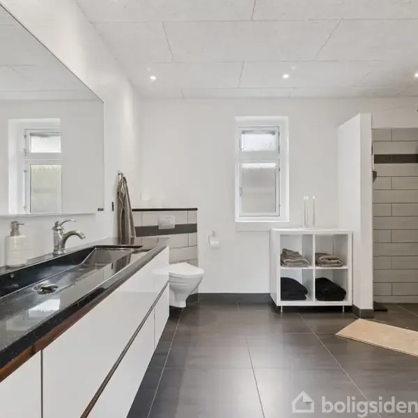 Dobbel håndvask med sorte bordplader og store spejle, brusekabine til højre med beige gulvmåtte, og hylde med foldede håndklæder i et lyst og rummeligt badeværelse.