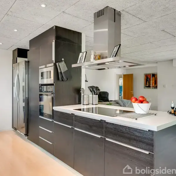 Et moderne køkken med sorte skabe og indbygget kogeplade. Ved siden af er der en stor, sølvfarvet køleskab. Bordpladen er dekoreret med en skål med røde æbler.