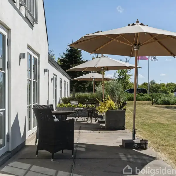 Terrassemøbler er arrangeret under store parasoller ved siden af et hvidt hus. I baggrunden ses en græsplæne med planter og træer under en blå himmel.