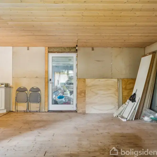 Et rum med ubehandlet trægulv; to sammenfoldede stole står ved en dør placeret i midten. Et køkkenmodul er til venstre, mens brædder læner mod højre væg.