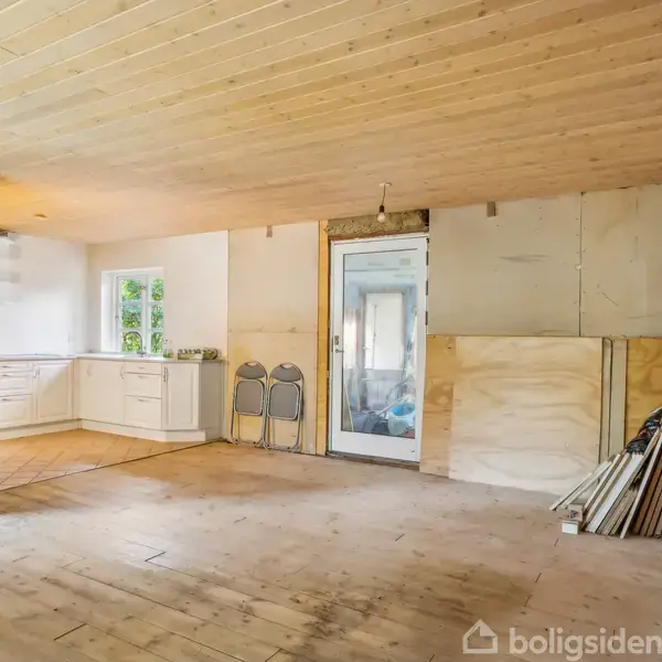 Et tomt rum med trægulv og træloft. En køkkensektion til venstre med hvide skabe. To klapstole står op ad en væg ved siden af en dør. Byggematerialer ligger i højre hjørne.