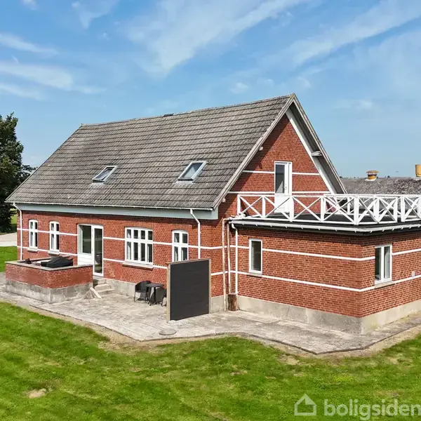 En rødstensbygning med skråt tag og tagvinduer står på en grusbelagt gårdsplads. Omkranset af en græsplæne og blå himmel, har huset en hvid dør og mange vinduer.