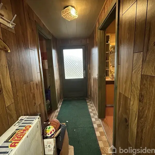 Træbeklædt gang fører til en glasdør, med grøn løber på gulvet. Til venstre ses en stigehylde med farverige bolde. Værelser ligger på begge sider af gangen.