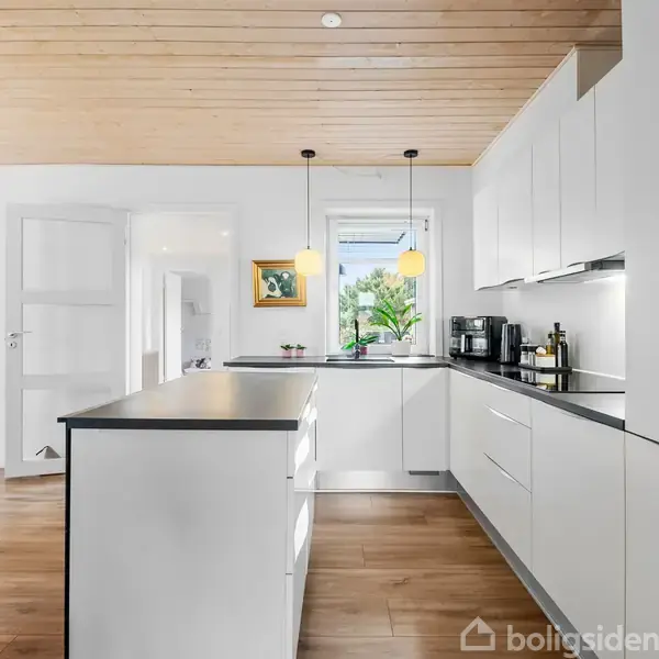 Et moderne køkken med hvide skabe og mørk bordplade. To pendellamper hænger over køkkenøen. Et vindue med grøn udsigt findes i baggrunden. Trægulv og træloft skaber en varm atmosfære.