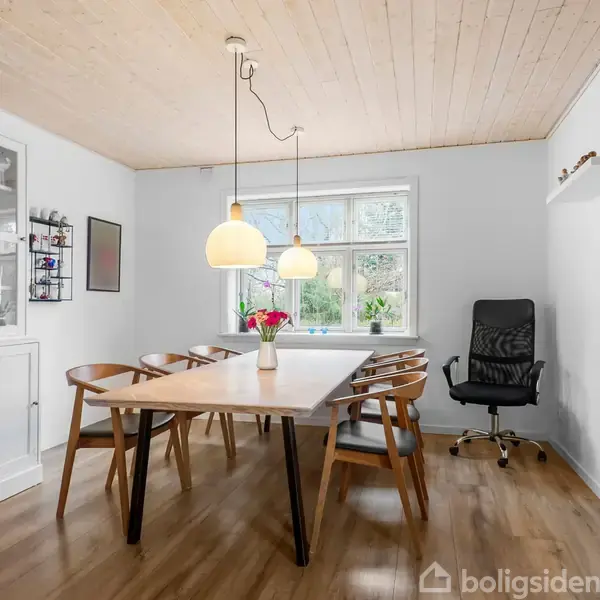 Et spisebord med seks stole står i et lyst rum med trægulv. To lamper hænger over bordet. En sort kontorstol er ved vinduet, og et hvidt skab står til venstre.