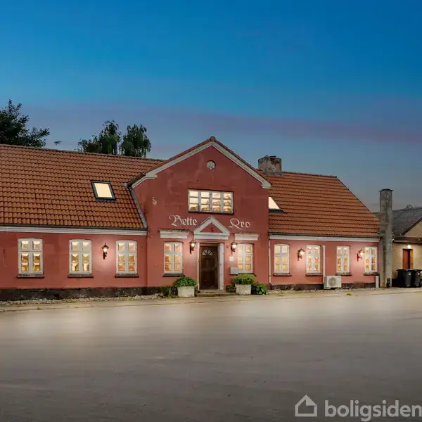 En rød bygning med et skilt, hvor der står "Motte Kro", oplyst mod en blå aftenhimmel. Bygningen har mange vinduer og ligger ved en vej.