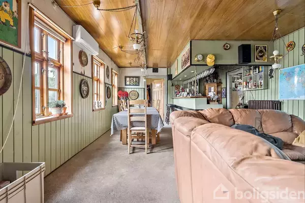 En stue med beige sofa i forgrunden, spisebord til seks personer, diverse dekorationer på grønne træpanelvægge og et hjørnebarområde. Ljuligt og traditionelt interiør med træloft.