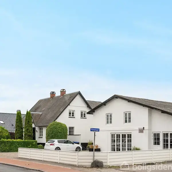 To hvide huse står langs en vej. En hvid bil er parkeret i indkørslen. Omkring er en hvid stakit og grønne buske. Vejskilt viser "Hvidovrevej."